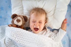 picture of a screaming baby lying in their bed with a teddy bear