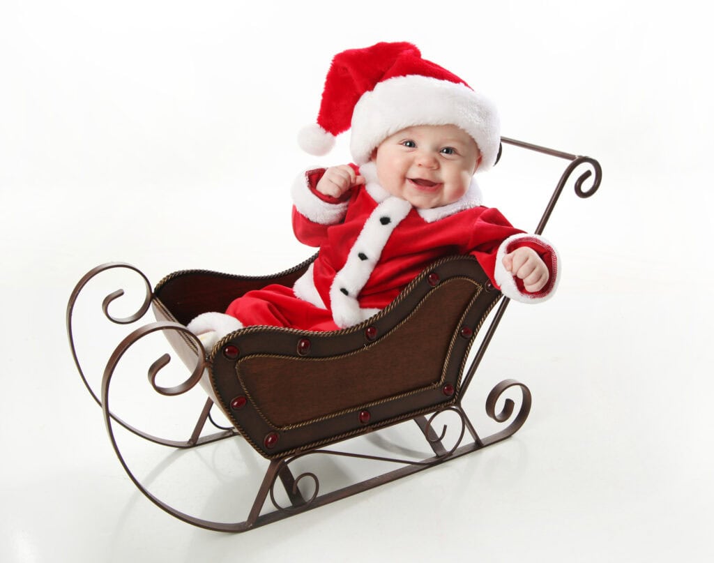 Adorable young baby wearing a santa claus suit and hat sitting in a metal Christmas snow sleigh