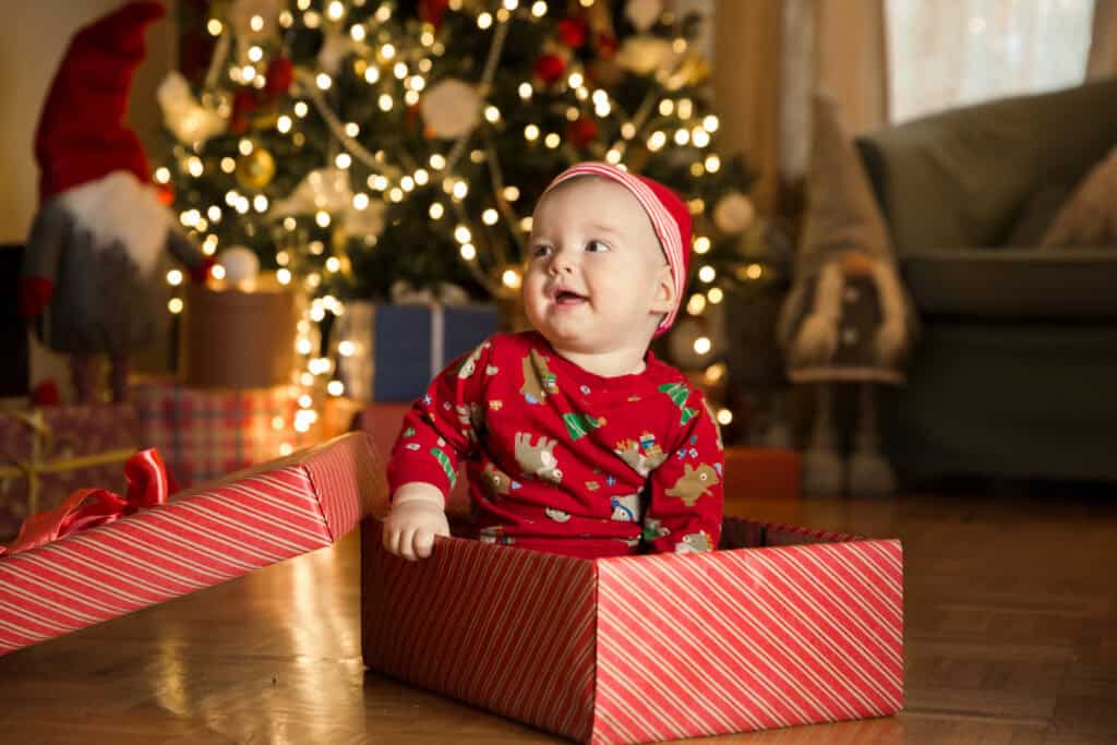 picture of a smiling baby wearing a Christmas outfit sitting in a Christmas gift box in front of a Christmas tree