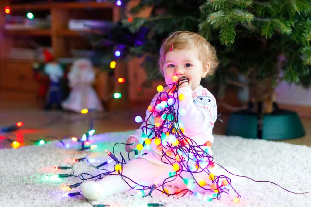 picture of a baby sitting in front of a Christmas tree chewing on a strand of Christmas lights