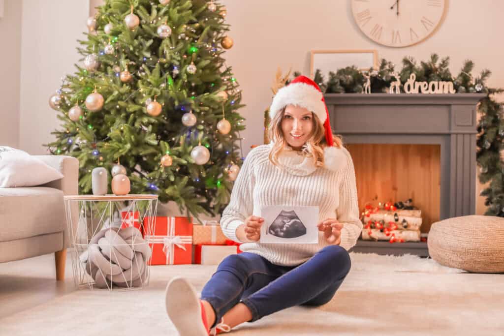 pregnant woman sitting in front of a Christmas tree while holding an ultrasound picture