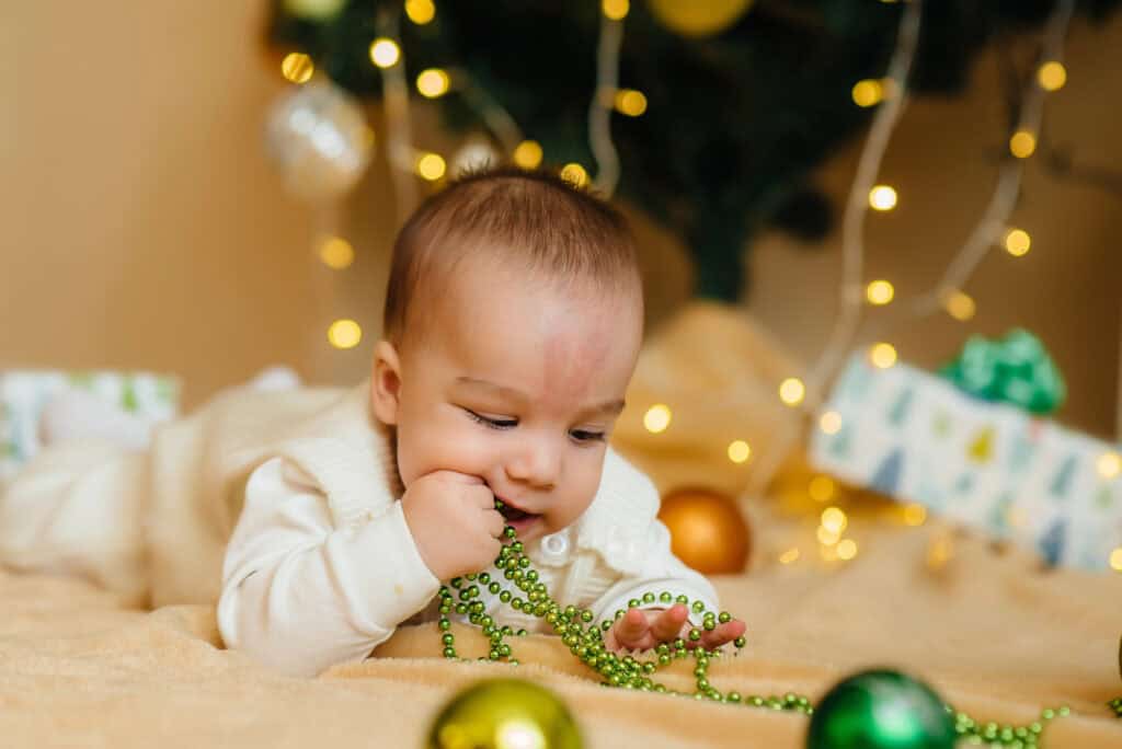 small baby lying under a Christmas tree chewing on a stand of Christmas lights