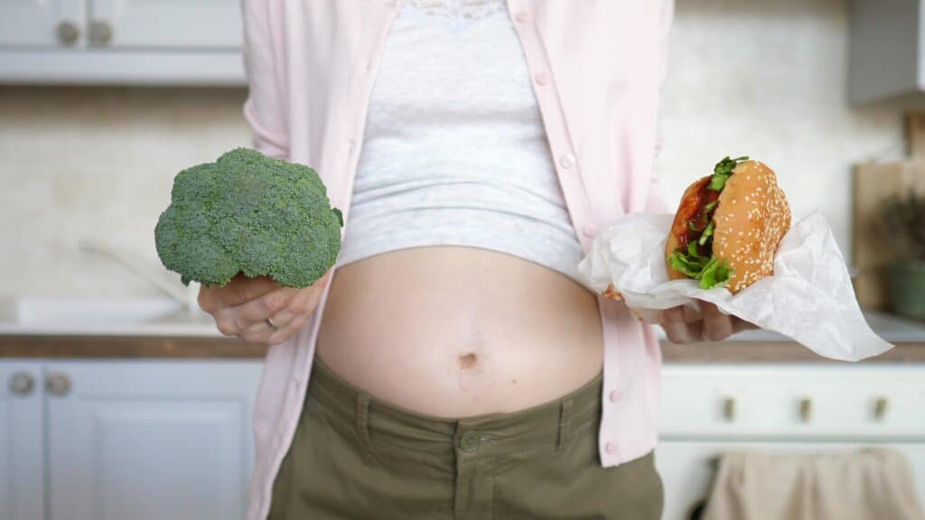 picture of a pregnant woman holding broccoli in one hand and a juicy cheeseburger in the other hand