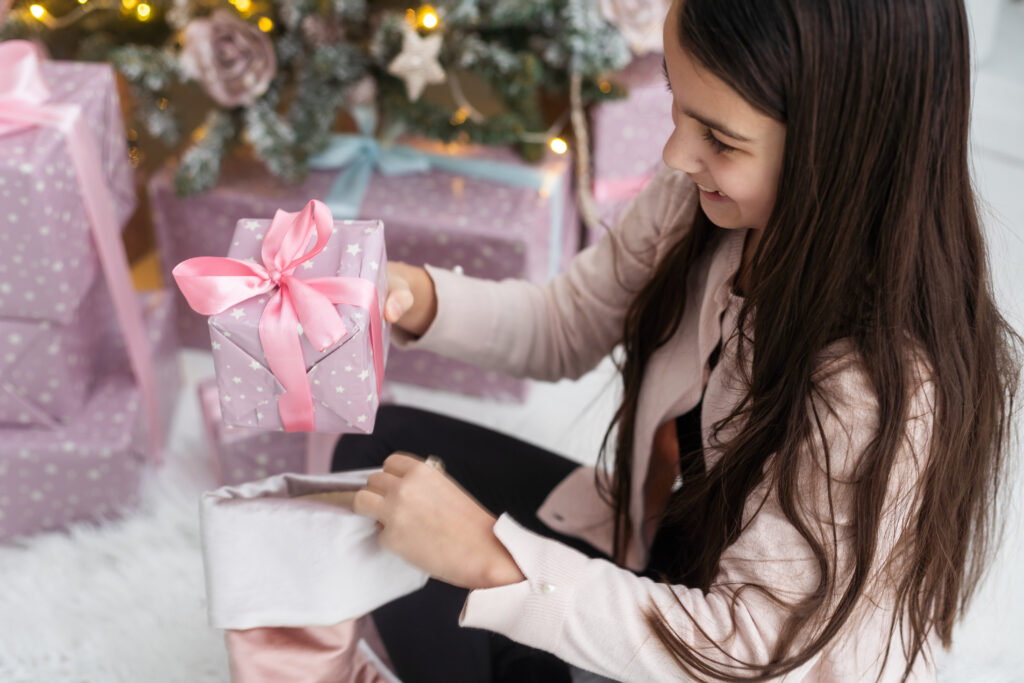 picture of a tween girl sitting beside a Christmas tree with many wrapped Christmas gifts