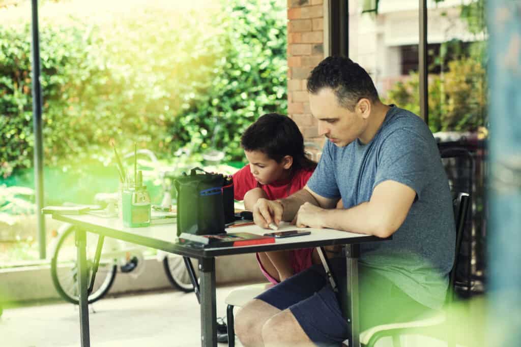 father sitting on the back porch at a table doing homeschool work with his child