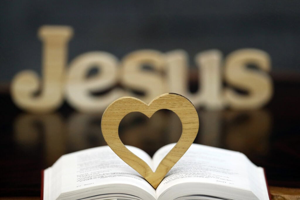 picture of a Bible laying open with a wooden heart sitting up in the middle with the word JESUS in the background