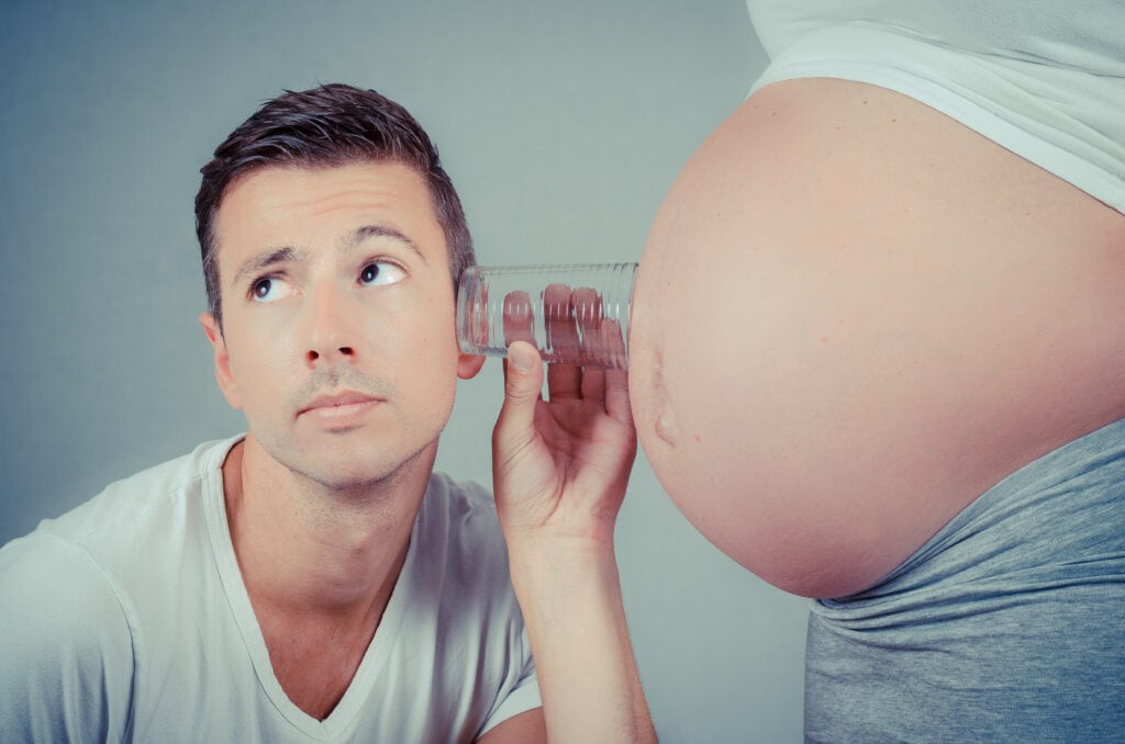 picture of a man holding a glass to his pregnant wife's belly pretending to hear the baby inside